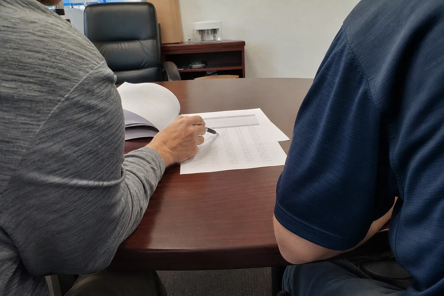 Over-the-shoulder view of two men at a wooden conference table, one pointing a pen at a printed page of figures in a small office