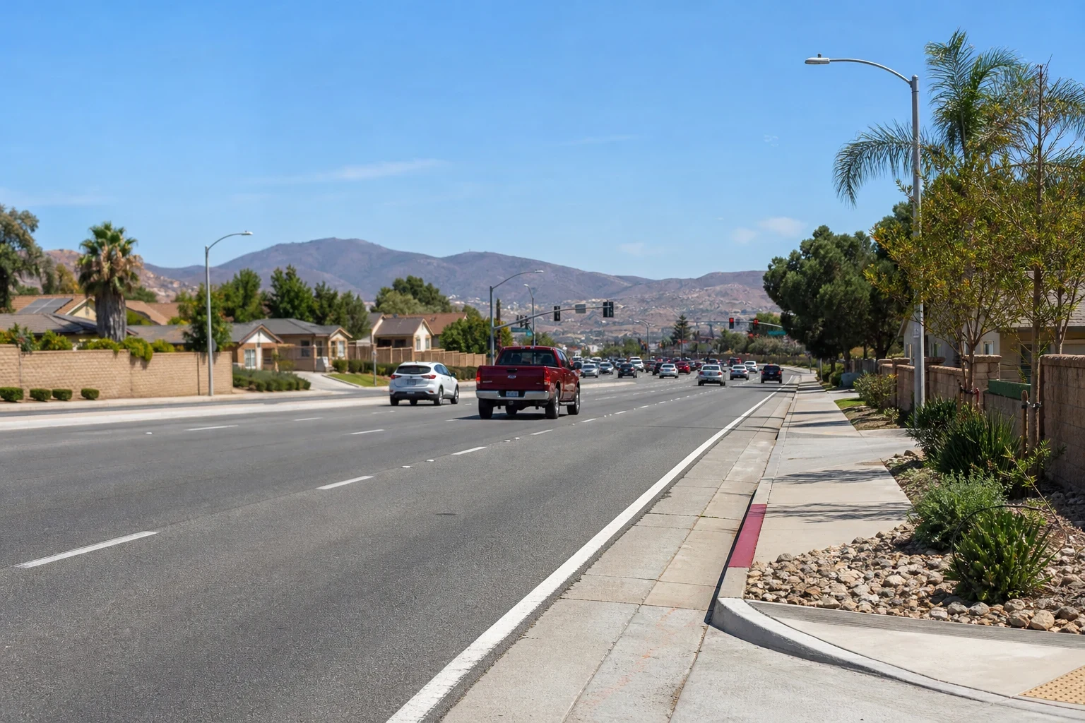 Wide multi-lane suburban road with light traffic, a red pickup, block walls, palm trees, tile-roof homes and brown hills under a clear blue sky