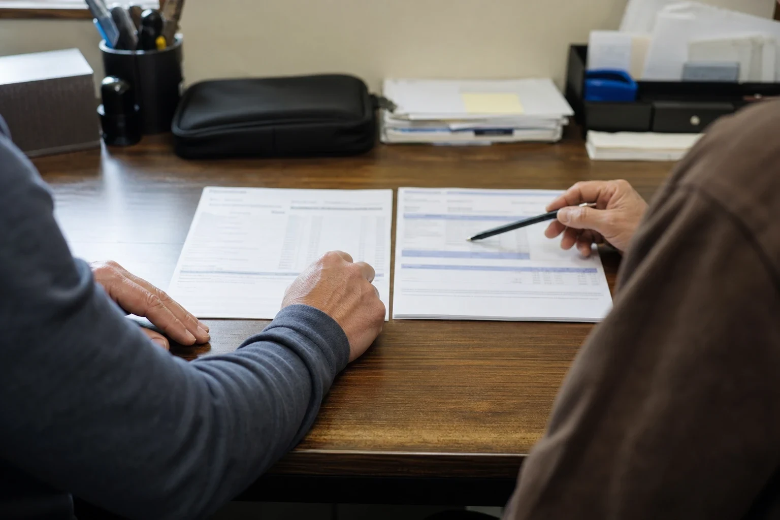 Two people seated at a wooden office desk, one pointing a pen at a document laid beside another form, with a pen cup, black pouch and paper trays behind