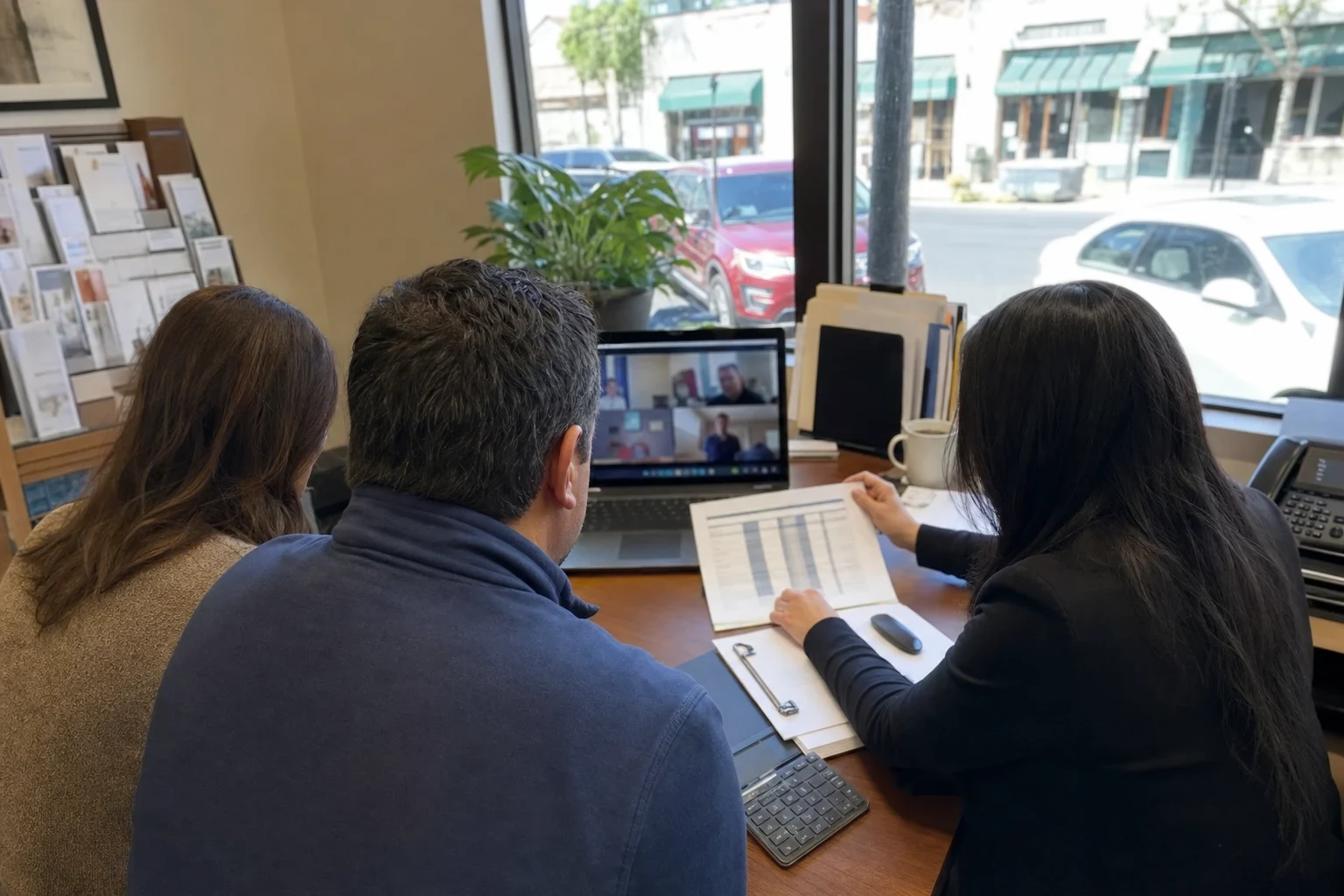Agent at an office desk by a street-facing window reviews a printed chart with a couple, with a laptop, folders, coffee mug and brochure rack nearby